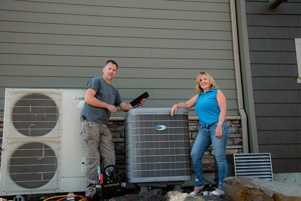 HVAC technician and woman posing beside residential air conditioning unit and Mitsubishi heat pump system outside home.