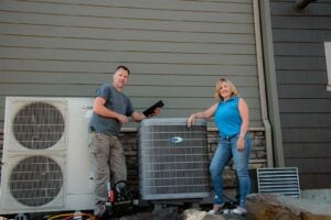 HVAC technician and woman posing beside residential air conditioning unit and Mitsubishi heat pump system outside home.