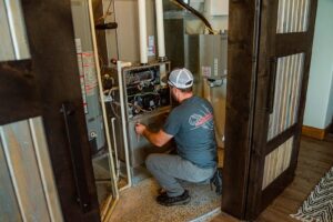 Cougar technician in white cap crouches to inspect interior components of a residential furnace unit during maintenance.