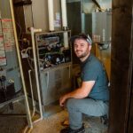 HVAC technician crouching beside an open furnace unit during installation or maintenance work in a residential home.