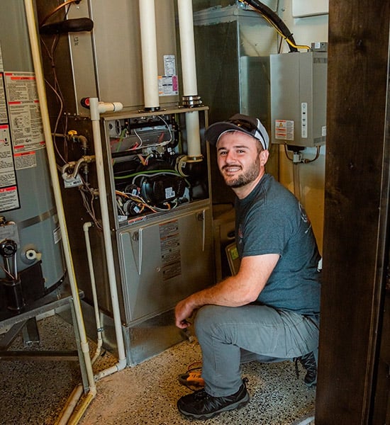 HVAC technician crouching beside an open furnace unit during installation or maintenance work in a residential home.