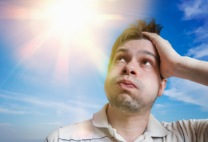 Man looking worried and overheated under bright sun against blue sky, scratching his head in distress.