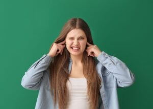 Young woman with long brown hair wearing a light blue denim jacket and white top, smiling broadly with hands near her ears against a green background.