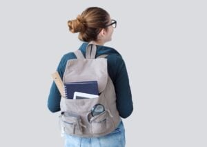 Student wearing glasses and a beige backpack filled with notebooks, spiral-bound notebook, and school materials stands against a white background.