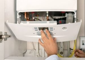 Technician's hand adjusting temperature dial on white boiler control panel with visible copper pipes and internal components above.