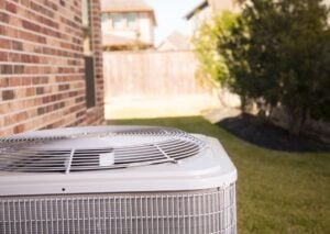 Air conditioning unit installed in residential backyard with brick home, green lawn, and hedges visible in background.