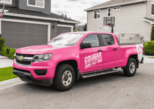 Bright pink Chevrolet Colorado pickup truck branded with Cougar Heating and Air Conditioning logo and phone number (509) 342-7050 parked in a residential drivew