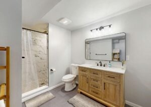 Spacious modern bathroom featuring a white toilet, wooden vanity with sink, framed mirror, and walk-in shower with marble tile.