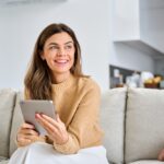 Woman sitting on couch holding tablet while reading service information with satisfied expression.
