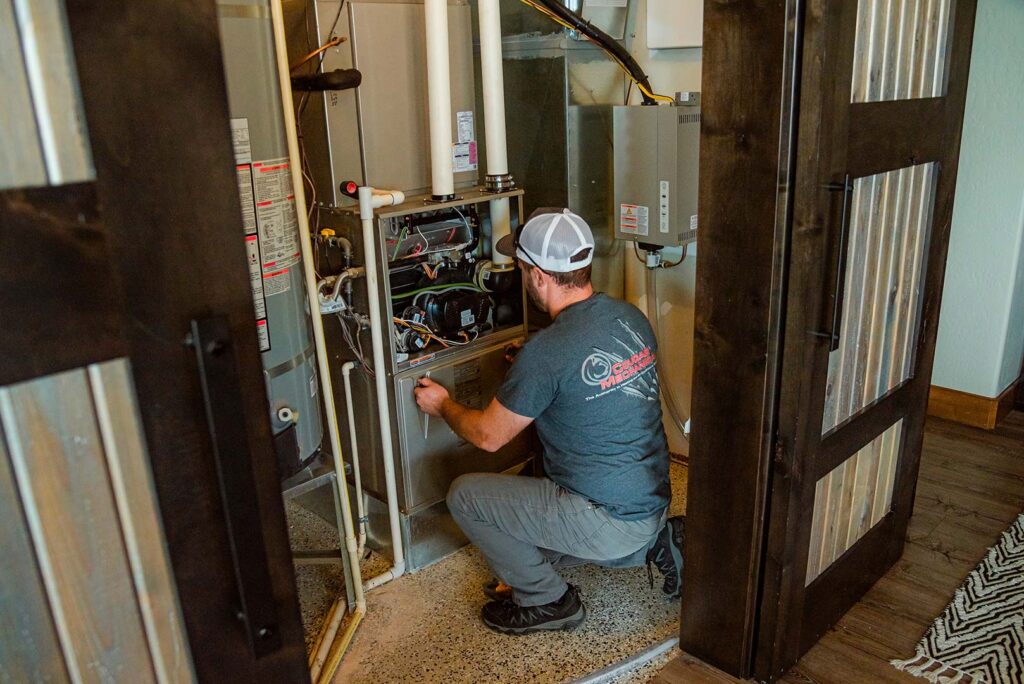 Cougar technician in white cap crouches to inspect interior components of residential furnace unit in utility space.